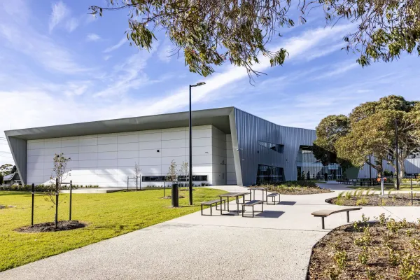 Norlane Arc Exterior view of a modern community building with light grey and dark metal cladding, set beside a landscaped lawn. A paved pathway with benches and small trees leads to the entrance under a blue sky.