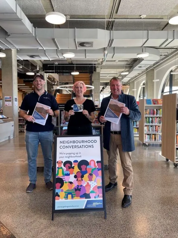 Cr Chris Burson, Cr Emma Sinclair and Cr Ron Nelson at a Neighbourhood Conversations event Three people standing indoors behind a freestanding sign that reads ‘Neighbourhood Conversations – We’re popping up in your neighbourhood’. Each person is holding an information flyer. Bookshelves and seating are visible in the background, indicating a public library or community space.