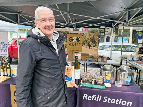 Steve Parker of Geelong Region Olives Steve Parker is a man with white hair and glasses and he stands in front of his market stall wearing a black raincoat and with one hand is holding a bottle and can of olive oil.