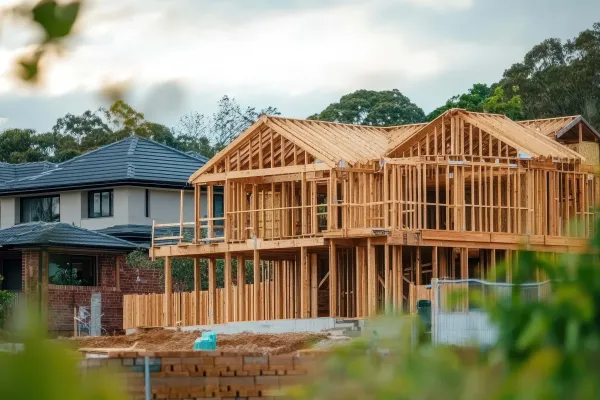 Stock building construction Two‑storey house under construction with exposed timber framing, next to completed homes and surrounded by trees.