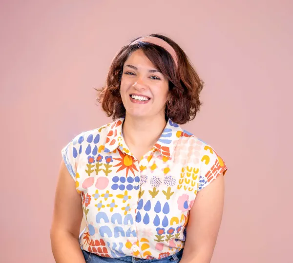 Hadeel Image of young woman sitting in front of pink background with pink headband and brightly coloured patterned shirt