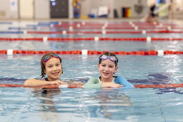Swimmers enjoying the City's Swim Sport & Leisure facilities. Two young swimmers with goggles on their heads are in the pool leaning on a lane rope and smiling.