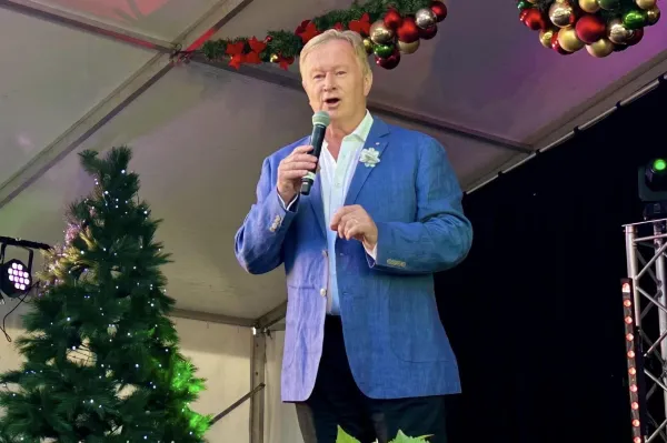 Denis Walter "Australian entertainer Denis Walter speaks into a microphone on an outdoor stage decorated with Christmas trees and festive baubles."